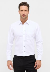 Hvid langærmet skjorte med almindelig krave, der har sorte knapper og en lomme på venstre side, parret med sorte bukser.