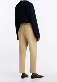 Maglione navy a trama, pantaloni beige su misura con vestibilità dritta e scarpe nere basse. I pantaloni presentano tasche posteriori e accenti sottili.