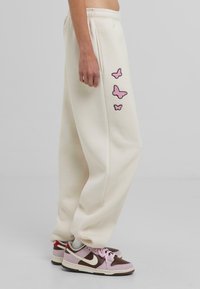 Crèmekleurige sweatpants met roze geborduurde vlinderontwerpen aan de zijkant. Het model draagt bruin en roze sneakers.