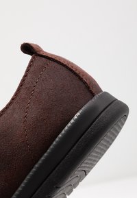 Hnědá semišová nazouvací bota s kulatou špičkou, měkkou texturou a černou gumovou podrážkou se vzorovaným protiskluzovým povrchem.