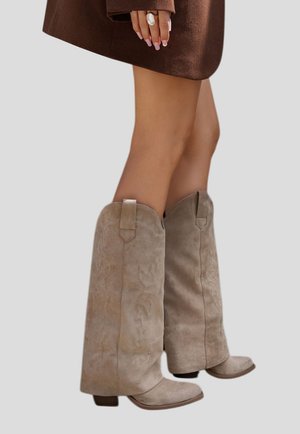 Jambes dans des bottes de cowboy en daim beige, mi-mollet, associées à une jupe marron, main reposant sur la cuisse avec une grande bague blanche.