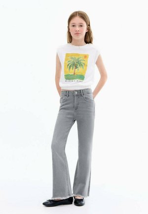Weißes ärmelloses Hemd mit Palmenbaum-Graphic; graue Schlagjeans mit ausgefransten Säumen; schwarze Schuhe. Das Modell steht mit Händen in den Hüften.