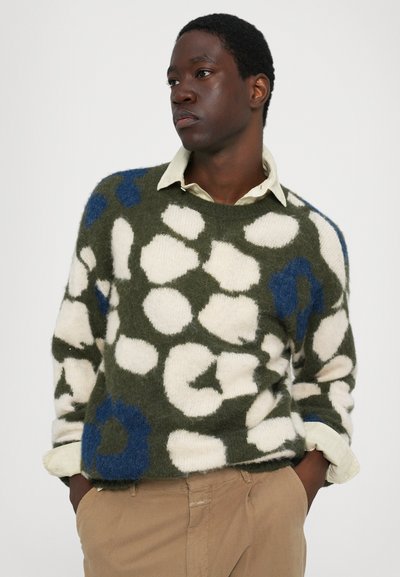 Suéter verde con patrón blanco y acentos azules, hecho de material suave. Cuello redondo, corte relajado, superpuesto a una camisa clara.