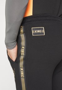 Černé joggingové kalhoty s gold logem "KING" a pruhy. Elastický pas, boční kapsy. Prodyšný materiál, vypasovaný střih.