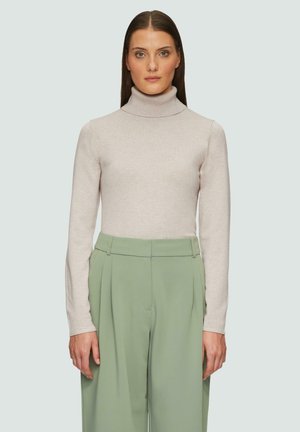 Un pull à col roulé beige clair avec des manches longues, en texture tricot. Associé à un pantalon vert taille haute, ample.