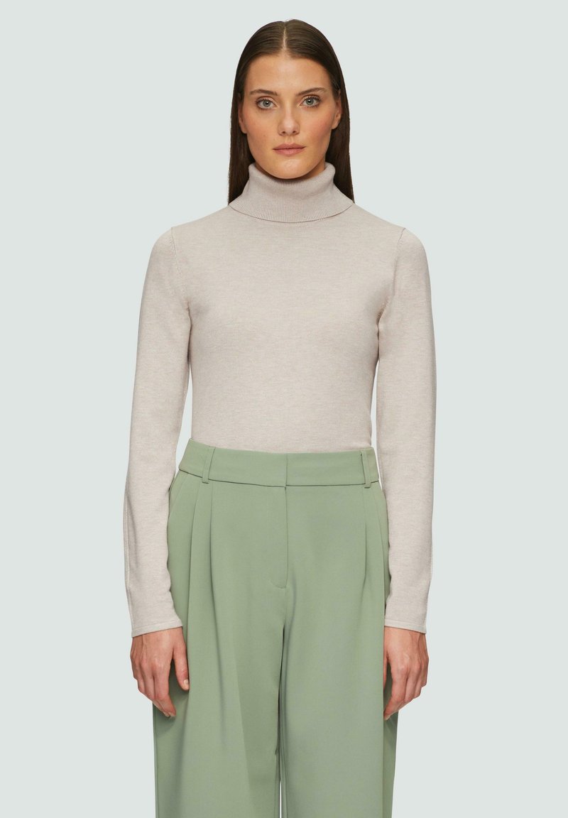 Hellbeige Rollkragenpullover mit langen Ärmeln und gestrickter Textur. Kombiniert mit hochgeschnittenen, locker sitzenden grünen Hosen.