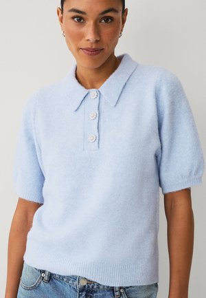 Vrouw draagt lichtblauw kort mouwsweater met kraag en drie decoratieve knopen, gecombineerd met blauwe spijkerbroek.