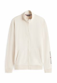 Veste zippée beige clair avec un col haut, des poches avant et de subtils accents de logo sur la manche. Texture de tissu lisse.