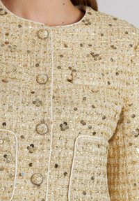 Gold- und cremefarbener Tweedjacke mit runden Pailletten, runden Knöpfen und kontrastierenden Paspeln entlang der Kanten. Strukturierter Stoff mit einem strukturierten Design.