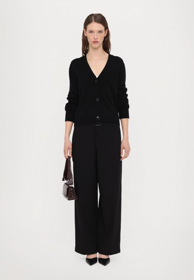 Black knit cardigan with a V-neck, front buttons, and ribbed cuffs, paired with wide-leg black trousers and a small dark handbag.