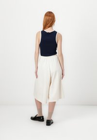 Tamno plava bez rukava s bijelim rubom, uparena s kremastim culottes hlačama. Model nosi crne cipele i bež čarape. Pogled sa stražnje strane na bijeloj pozadini.