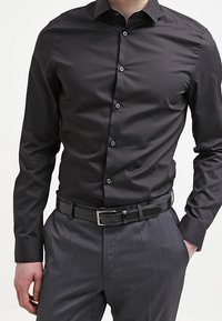 Camicia nera in cotone con colletto a punta, maniche lunghe e chiusura con bottoni, abbinata a pantaloni grigi e una cintura in pelle nera.