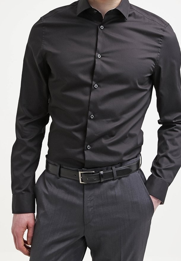 Camicia nera in cotone con colletto a punta, maniche lunghe e chiusura con bottoni, abbinata a pantaloni grigi e una cintura in pelle nera.