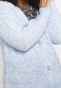 Hellblauer gestrickter Cardigan mit strukturiertem Muster, langen Ärmeln und Perlmuttknopfdetails. Weiches Garn mit gemütlichem, entspanntem Design.