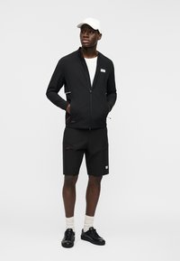 Homme portant une veste à fermeture éclair noire, un short noir, une casquette blanche, des chaussettes blanches et des baskets noires, debout les mains dans les poches, regardant vers la droite.