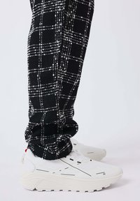 Pantaloni a scacchi bianchi e neri inseriti in sneakers sportive bianche con suole spesse e dettagli neri minimi.