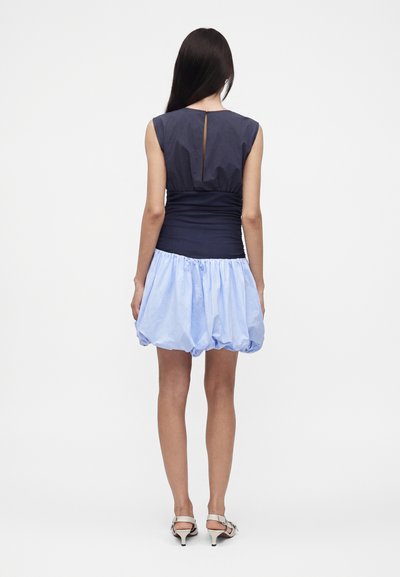 Femme aux longs cheveux foncés portant un haut sans manches bleu marine et une mini-jupe bleu clair bouffante, debout de dos avec des talons blancs.
