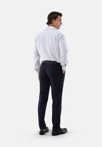 Camicia a maniche lunghe a quadri bianca e blu, pantaloni scuri, cintura nera e scarpe; posizione in piedi, vista da dietro.