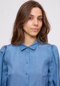 Camicia blu chiaro con abbottonatura frontale, colletto a punta e maniche a sbuffo. Realizzata in tessuto liscio e lucido con una sottile texture.
