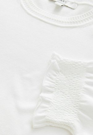 Vit tröja med rund halsringning, texturerade volangdetaljer på ena sidan och slätt tyg över hela. Etikett synlig på toppen.
