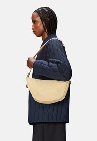 Borsa a spalla in pelle gialla con un design curvo, tenuta da una persona che indossa una giacca trapuntata blu navy. La tracolla presenta un accento hardware discreto.