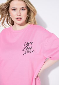 Różowa bawełniana koszulka z krótkim rękawem, zdobiona czarnym graficznym wzorem z napisem "Love" powtarzającym się w różnych stylach na przodzie. Luźny krój.