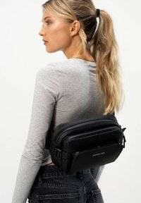 Marsupio nero in nylon con più scomparti zip, texture liscia, tracolla regolabile, indossato sulla schiena. L'utente indossa una maglietta grigia.