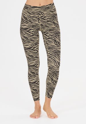 Ben iført højtaljede, beige leggings med sorte zebrastriber, stående barfodet mod en ensfarvet lys baggrund.