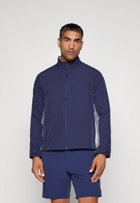 Marineblå hettejakke med grå detaljer, laget av lett og elastisk stoff. Har høy krage og frontlommer. Shorts i matchende marineblå.
