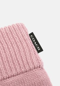 Rosa stickad accessoar med ribbad textur och en tydlig svart etikett med "COACH" i vit text.