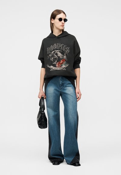 Γυναίκα που φορά στρογγυλά μαύρα γυαλιά ηλίου, μαύρη μπλούζα με κουκούλα με σχέδιο πάνθηρα, τζιν παντελόνι με πλατιά πόδια δύο τόνων, μαύρα παπούτσια και κρατά μια μαύρη τσάντα.