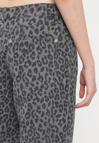 Jeans s leopardím vzorom v šedej farbe s textúrovanou látkou. Obsahujú dve zadné vrecká a kovové nitovanie, zobrazené zo strany s odloženou rukou.