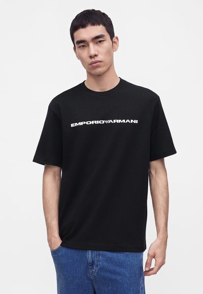 Νέος άνδρας που φοράει μαύρο T-shirt Emporio Armani και μπλε τζιν, στέκεται με το ένα χέρι στην τσέπη μπροστά σε απλό ανοιχτό φόντο.