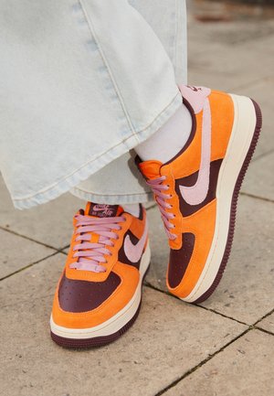 Persoon draagt oranje en bruine Nike Air Force 1 sneakers met roze veters en lichtgekleurde broek, staand op een stenen trottoir.