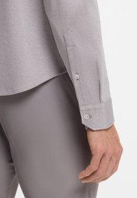 Ανοιχτό γκρι πουκάμισο με υφή υφάσματος, διαθέτοντας κανονικό γιακά και ρυθμιζόμενες μανσέτες με κουμπιά, συνδυασμένο με παντελόνι ίδιου χρώματος.