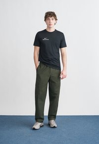 Crna pamučna majica s bijelim Nike logotipom, uparena s tamnozelenim cargo hlačama i sivim tenisicama. Model stoji na neutralnoj pozadini.