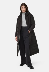 Zwarte geïsoleerde lange jas met een capuchon, met een doorgestikte textuur en ritssluitingen aan de zijkant, gedragen over een grijze coltrui en brede broekspijpen.