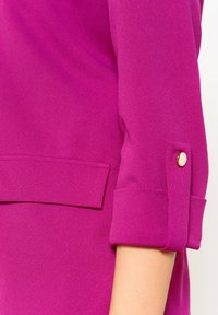 Blusa fucsia con una texture liscia, caratterizzata da maniche arrotolate e un dettaglio con bottone dorato sul polsino. Design sartoriale con un taglio delicato.