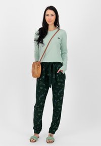 Lichtgroen overhemd met lange mouwen, donkergroene broek met trekkoord en bloemenpatroon, beige ronde tas en mintgroene slip-on sandalen.