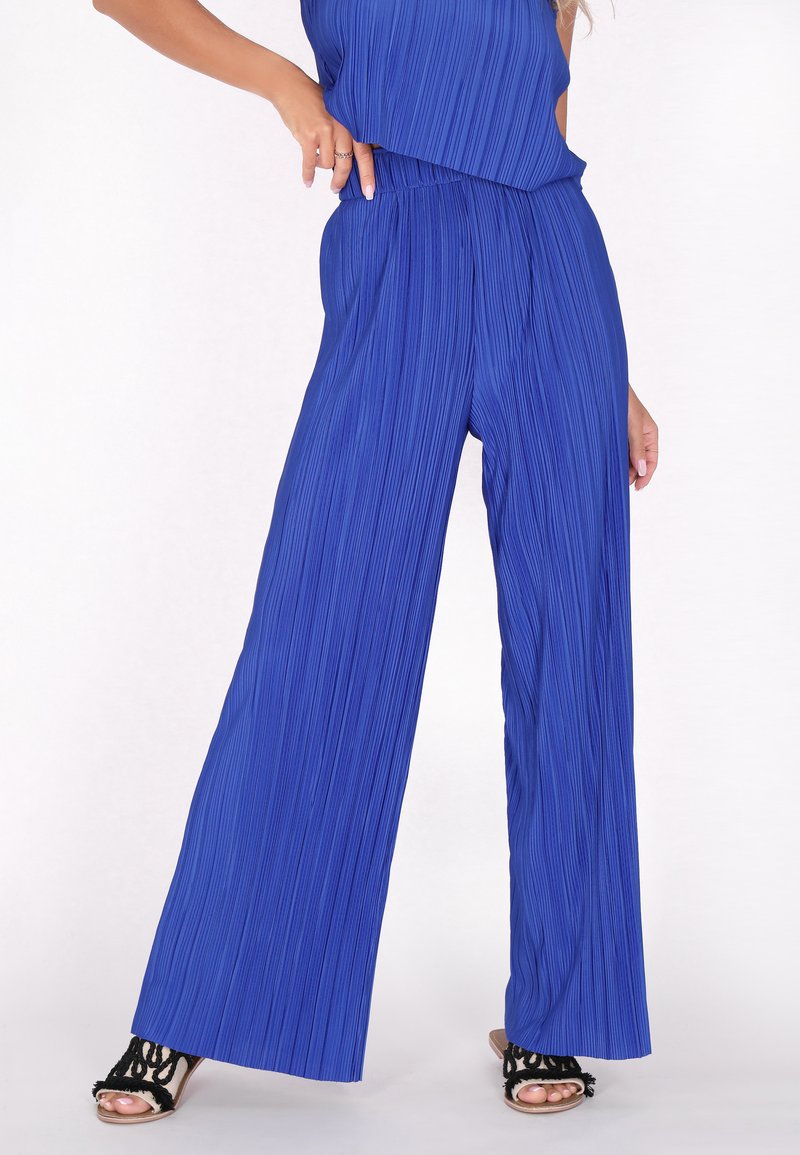 Blauwe, geplooide wijde broek met een elastische tailleband, gemaakt van een gestructureerde stof. Zwarte sandaaltjes met bandjes maken de outfit compleet.