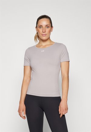 Egy nő áll, világosszürke, testhez simuló, rövid ujjú sportpólót és fekete leggingset viselve, egy sima fehér háttér előtt.