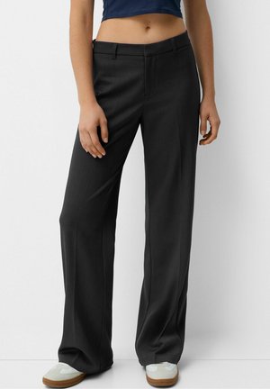 Kvinde iført sorte bukser med vide ben, hvide sneakers og en marineblå cropped top, stående mod en ensfarvet hvid baggrund.