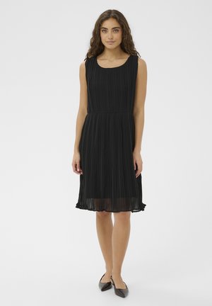 Kobieta stojąca, ubrana w czarną plisowaną sukienkę bez rękawów do kolan oraz czarne, spiczaste baleriny, na jednolitym białym tle.