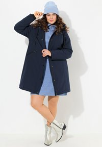 Navy hooded coat with a zippered pocket, worn over a light blue ribbed dress and silver ankle boots. The coat has black accents and a relaxed fit.