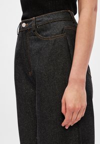 Jans pretos com uma textura de denim, apresentando um design de cintura alta, costura laranja e um fecho de botão prateado.