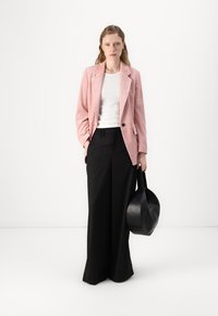 Blazer en velours côtelé rose avec revers en angle, associé à un top blanc à côtes et un pantalon noir à large jambe. Le mannequin tient un sac rond noir.