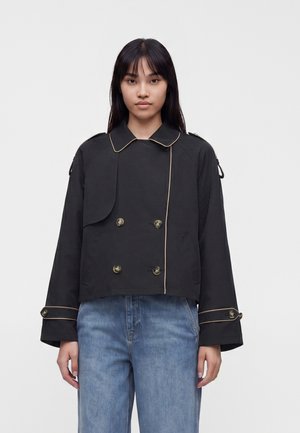 Jeune femme aux longs cheveux noirs porte une veste courte croisée noire avec un passepoil doré et un jean bleu sur un fond blanc uni.