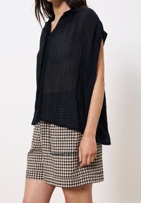 Schwarzes, strukturiertes, kurzärmliges Oberteil mit Kragen und Knopfleiste; kombiniert mit beige-schwarzen Karo-Shorts mit Taschen.