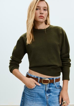 Frau mit langen blonden Haaren, die einen olivgrünen Pullover, einen blauen Jeansrock mit braunem Gürtel und goldenen Schmuck trägt, posiert mit der Hand in der Tasche.