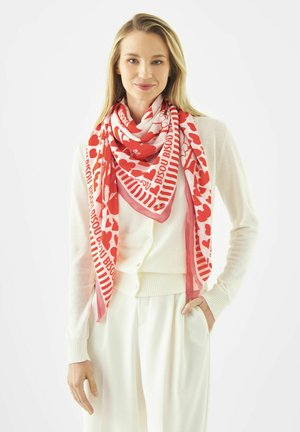 Vrouw draagt een witte cardigan en broek met een rood-wit sjaaltje met hartjes en tekst om haar nek gewikkeld.